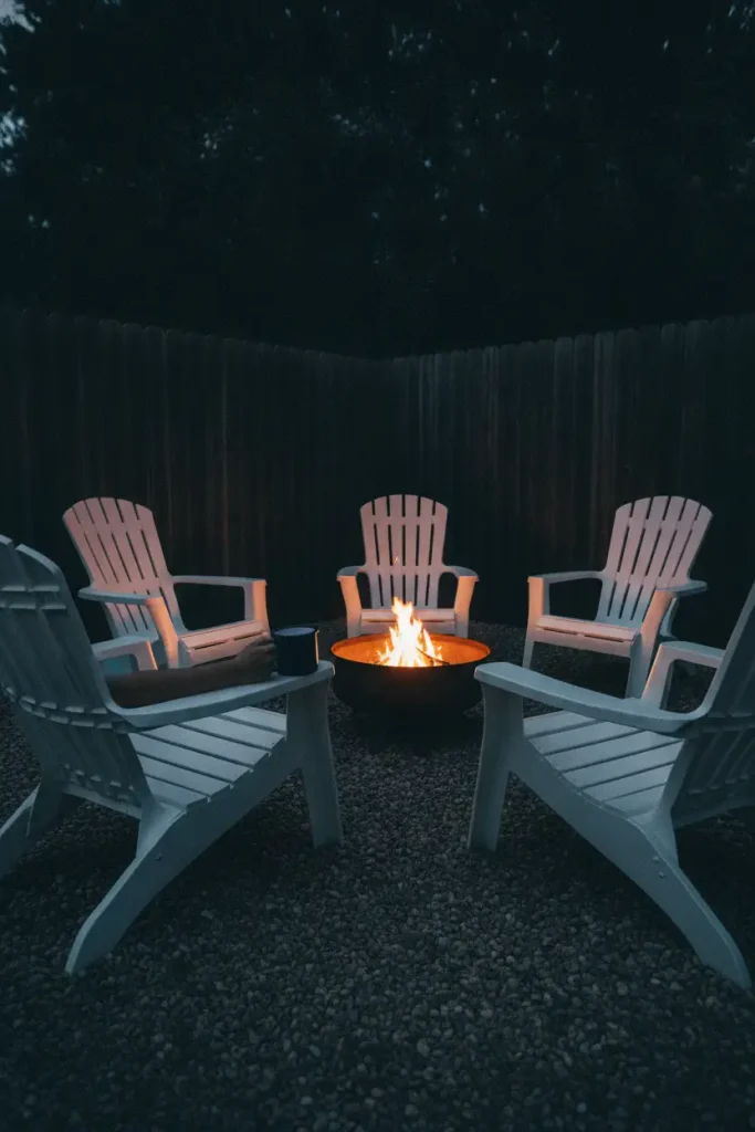 Corner fire pit ideas with white Adirondack chairs curved around a steel fire pit at night