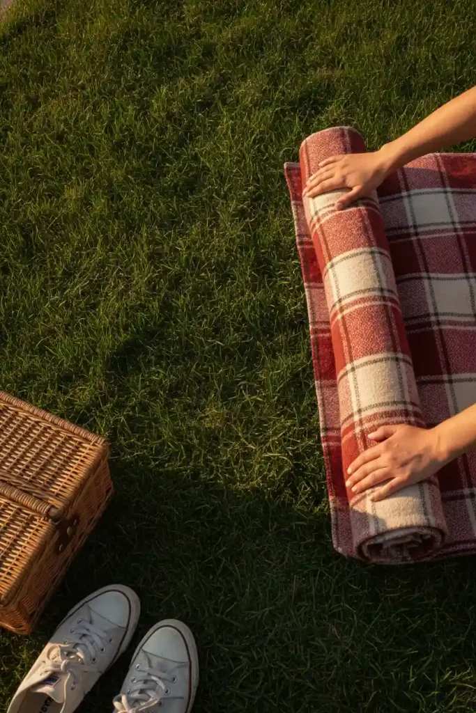 Hands rolling up a plaid picnic blanket from a green backyard lawn to protect the grass