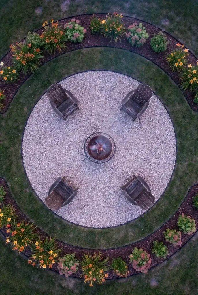 Overhead view of a fire pit with a gravel circle, chairs on the edge, and a daylily bed beyond the lawn